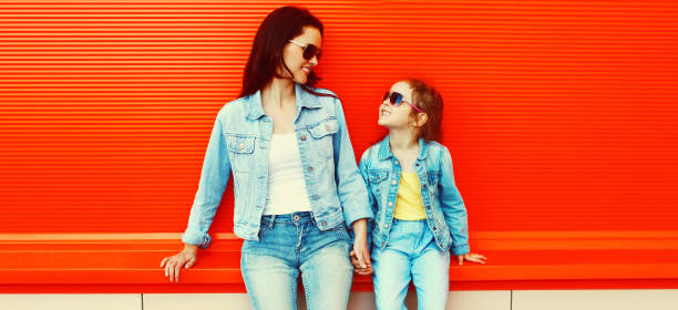 Portrait of stylish mother and child looking at each other wearing a sunglasses, denim clothes in a city on red background stock photo