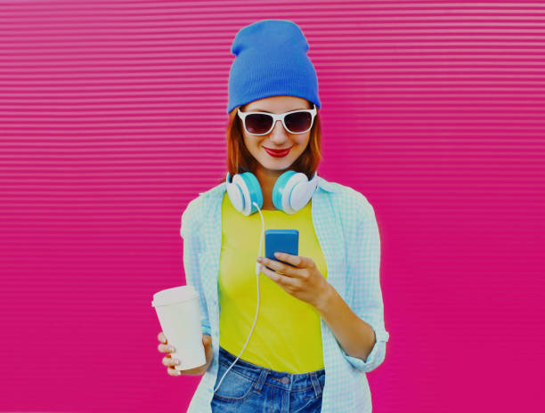 Portrait of stylish young woman with smartphone listening to music in headphones on pink background stock photo