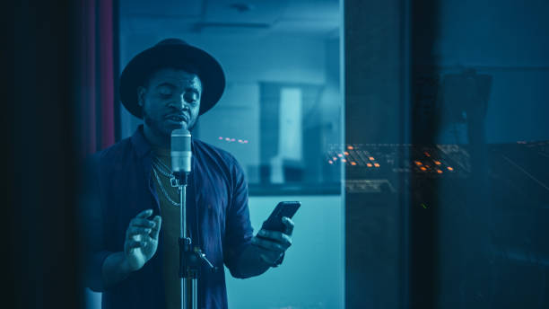 Portrait of Successful Young Black Artist, Singer, Performer Singing His Hit Song for the New Album. Wearing Stylish Hat, Holding Smartphone and Standing in Music Record Studio Soundproof Room.  rapper stock pictures, royalty-free photos & images