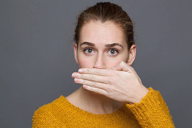 portrait of surprised young woman covering her mouth for silence negative feelings concept - portrait of surprised beautiful 20s girl covering her mouth for bad breath or taboo,studio shot on gray background bad breath stock pictures, royalty-free photos & images