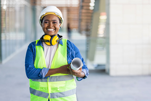 Portrait of woman engineer at building site looking at camera with copy space. Construction manager standing in yellow safety vest and white hardhat with crossed arms. Successful confident architect at construction site
