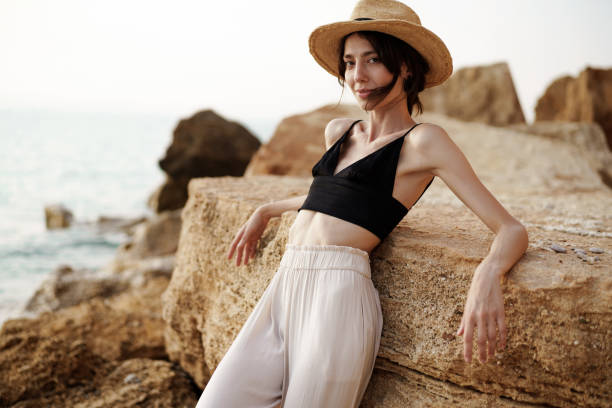 Portrait of woman in black bralette and white trousers leaning on rock at beach stock photo