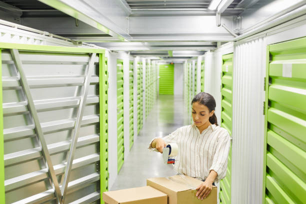 Portrait of Woman Packing Boxes in Storage Unit Wide angle portrait of young woman packing boxes with tape gun while standing by self storage unit, copy space self storage stock pictures, royalty-free photos & images