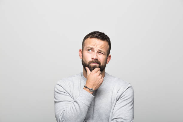 Portrait of worried bearded young man looking up with hand on chin Portrait of worried bearded young man looking up with hand on chin. Studio shot, grey background. asking stock pictures, royalty-free photos & images