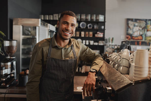 Portrait of young afro-american male business owner behind the counter of a coffee shop smiling looking at camera Handsome young male coffee shop owner standing behind counter barista stock pictures, royalty-free photos & images