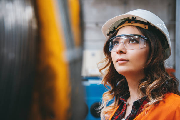 Portrait of young beautiful engineer woman working in factory building. Portrait of young businesswoman with white helmet looking up and seen from the industrial steel cable reel for crane in factory warehouse. She is testing and working with winding wire and cable drum accessories metal wire spool reel wear resistance. Engineer stock pictures, royalty-free photos & images