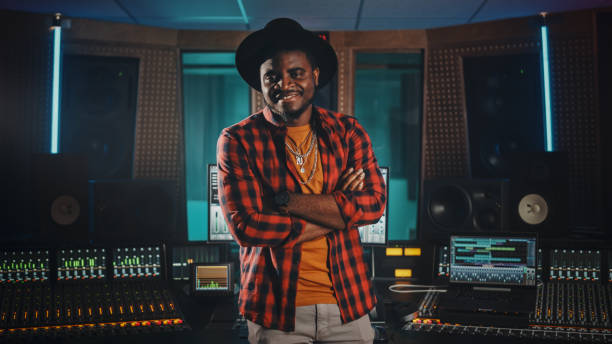 Portrait of Young Black Artist, Musician, Audio Engineer, Producer Wearing Stylish Hat and Standing in Music Record Studio. In the Background Control Desk, Mixing Equipment, Soundproof Room. Portrait of Young Black Artist, Musician, Audio Engineer, Producer Wearing Stylish Hat and Standing in Music Record Studio. In the Background Control Desk, Mixing Equipment, Soundproof Room. producer stock pictures, royalty-free photos & images