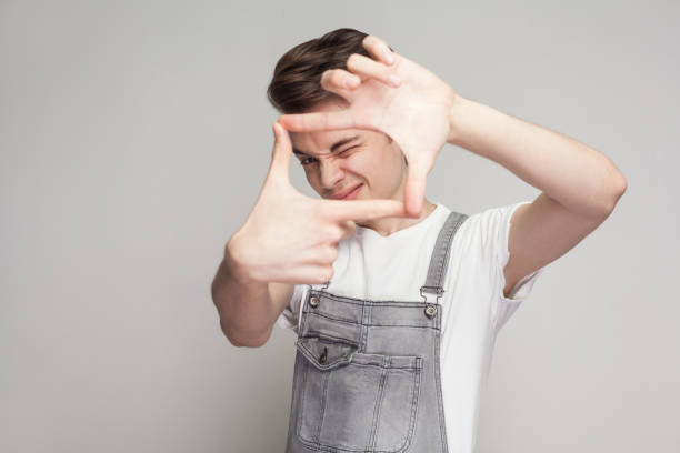 Portrait of young brunette man in casual style with white t-shirt and denim overalls standing and looking at camera and making camera frame with hands. Portrait of young brunette man in casual style with white t-shirt and denim overalls standing and looking at camera and making camera frame with hands. indoor studio shot, isolated on gray background. young male actors stock pictures, royalty-free photos & images