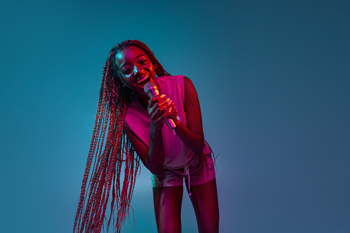 Singing, performing. Young joyful smiling artistic african woman singing with microphone isolated over blue background in neon lights. Concept of facial expression and human emotion.