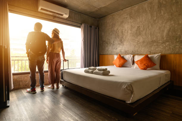 Portrait of young couple tourist standing nearly window, looking to beautiful view outside in hotel/resort bedroom after check-in. Conceptual of couple travel and vacation in their honeymoon period. airbnb stock pictures, royalty-free photos & images