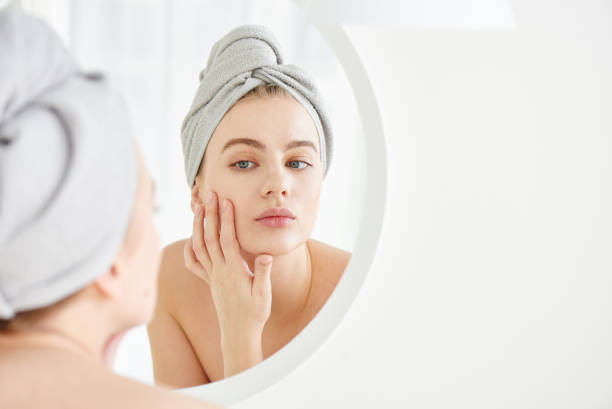 Portrait of  young girl with  towel on head in white bathroom looks and touches her face in the mirror and enjoys youth and hydration. Natural beauty, home care for problem skin stock photo