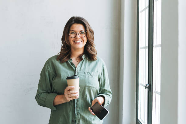 Portrait of Young smiling brunette woman plus size near window with paper cup of coffee and smartphone in hands in bright modern office stock photo