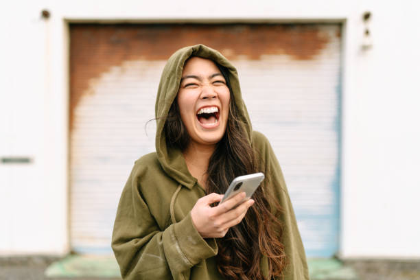 portrait of young woman holding smart phone and laughing - sorrir imagens e fotografias de stock
