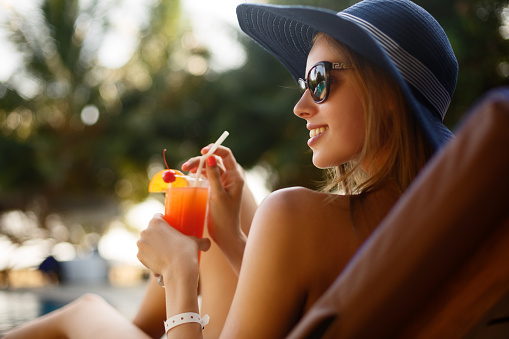 Portrait of young woman with cocktail glass chilling in the sun near swimming pool on a deck chair with palm trees behind. Vacation concept.