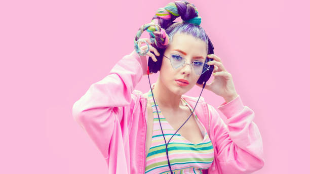 Portrait of young woman with headphones against pink background Confident fashionable funky young woman with creative hairstyle wearing cool hoodie standing against pink background and listening music on big headphones while looking at camera generation z stock pictures, royalty-free photos & images