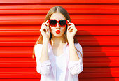 Portrait pretty young woman in red heart shape sunglasses over colorful background blowing lips