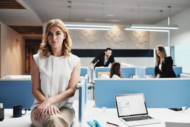 Portrait Worried Business Woman Looking At Camera In Coworking Office Portrait of serious female employee excluded by colleagues. Blonde businesswoman sitting on desk at work in modern coworking space, looking at camera with sad expression. exclusion stock pictures, royalty-free photos & images