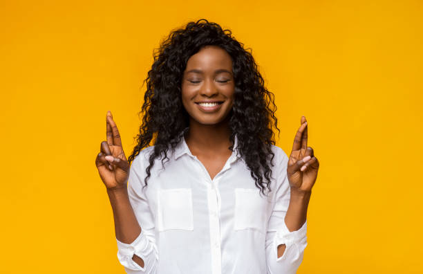 Positive african american girl crossing fingers, making wish Positive Black Woman Crossing Fingers With Closed Eyes, making wish, hopes for fortune and good luck, yellow background wanted signal stock pictures, royalty-free photos & images