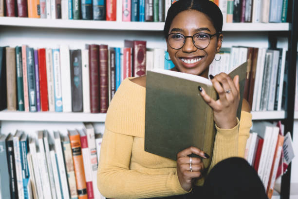 눈을 떼지 위해 안경을 쓴 긍정적인 아프리카계 미국인 젊은 여성이 책을 손에 들고 있는 동안 웃고 있습니다. 명랑 어두운 피부 학생 즐기는 문학 플롯 부터 베스트 셀러 - 서점 뉴스 사진 이미지