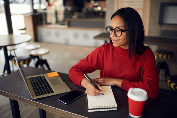 Positive dark skinned hipster girl in optical spectacles for vision correction looking away while creating new article with romantic idea in textbook for posting at own web blog via laptop device stock photo