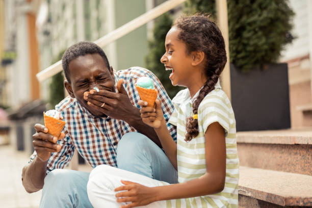 Positive delighted international male person eating ice-cream Creative father. Charming teenager laughing at funny situation, spending time with her daddy father and child stock pictures, royalty-free photos & images