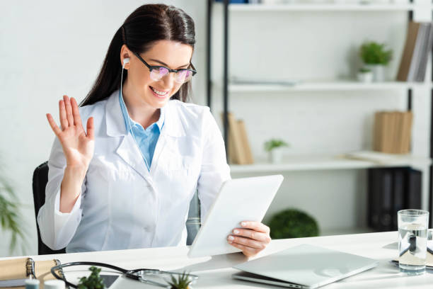 positive doctor waving and having online consultation on digital tablet in clinic office with laptop positive doctor waving and having online consultation on digital tablet in clinic office with laptop telemedicine stock pictures, royalty-free photos & images