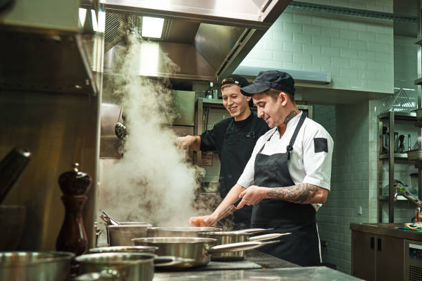 positive mood. side view of two cheerful and smiling cooks in uniform are preparing a food in a kitchen restaurant - chef imagens e fotografias de stock