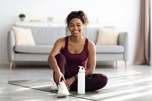 Positive sporty millennial black lady in beautiful sportswear and white sneakers sitting on fitness mat with bottle of water and smiling, having break while exercising at home, copy space