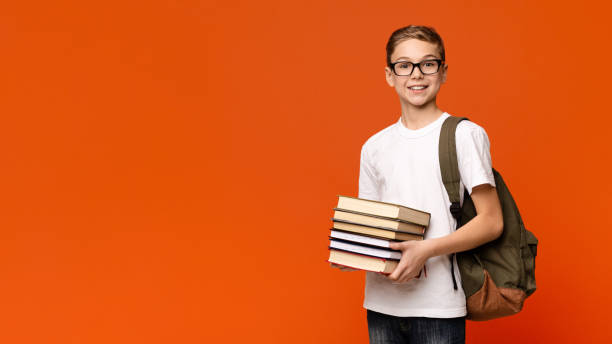 Positive teen boy in eyeglasses with backpack holding heap of books Loving knowledge. Positive teen boy in eyeglasses with backpack holding heap of books, orange panorama background with free space boys glasses stock pictures, royalty-free photos & images