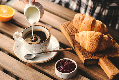 Pouring cream into cup of black coffee. Tasty breakfast table set with croissants, jam and coffee
