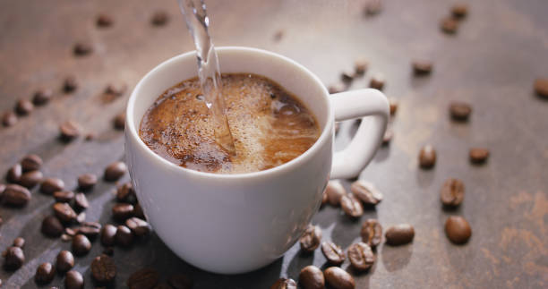 Pouring hot water into the cup of coffee and roasted coffee beans near the cup on the wooden table. Top view stock photo