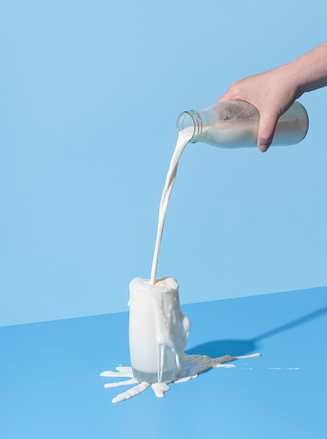 Woman hand pouring milk from a bottle into the glass, minimalist on a blue background. Over-spilling milk on the blue-colored table.