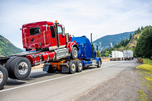 Powerful blue big rig semi truck transporting another semi truck tractor immersed by the front wheels on the frame of the first truck running on the road along the river stock photo