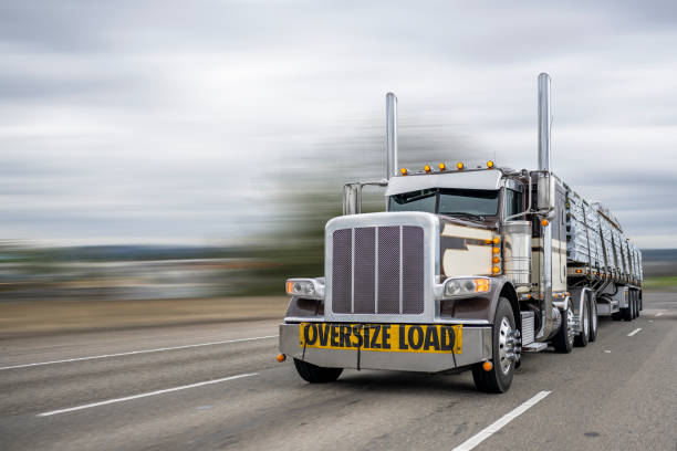Powerful classic dark big rig semi truck with oversize load sign on the front transporting lumber on the flat bed semi trailer running on the wide highway road stock photo