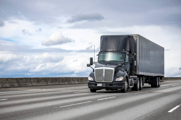 Powerful stylish black big rig day cab semi truck deliver commercial cargo in covered dry van semi trailer driving on the interstate highway road stock photo