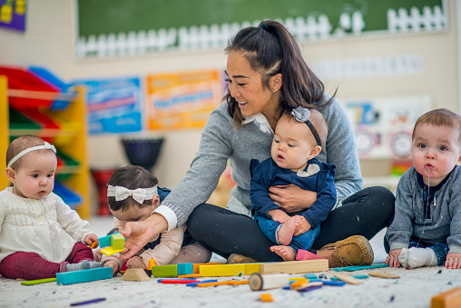 Smiling daycare teacher sitting and playing with a baby in her lap, with three other babies around her. 