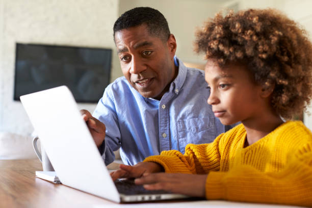 Pre teen girl black girl using a laptop computer sitting at table in the dining room with her home tutor, close up, selective focus  homework stock pictures, royalty-free photos & images