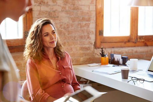 Pregnant businesswoman looking away at desk. Beautiful female professional is sitting in office. She is wearing shirt at workplace.