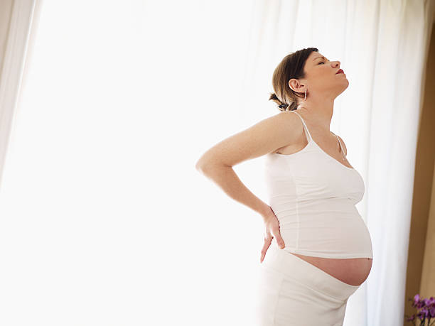 Pregnant woman holding back in front of white curtain stock photo