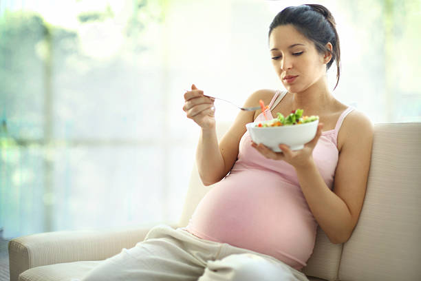 Pregnant woman relaxing at home and eating salad. Cute pregnant brunete woman relaxing on sofa and enjoying a vegetable salad.She's in late 20's.Wearing beige pregnancy pants and pink sleeveless tank top healthy diet pregnancy stock pictures, royalty-free photos & images