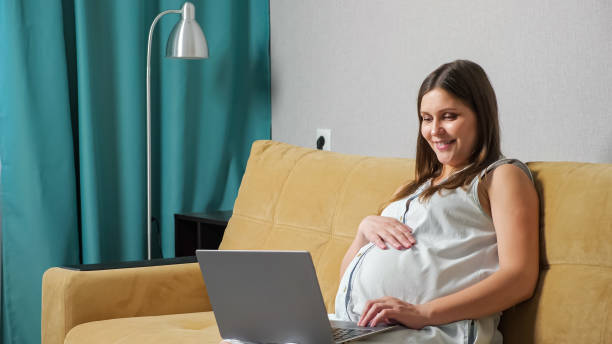 Pregnant woman talking on video call on laptop while sitting on sofa stock photo