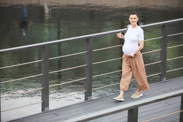 Pregnant woman walking along the bridge Portrait of young pregnant woman smiling at camera while drinking coffee and walking along the bridge outdoors walking during pregnancy stock pictures, royalty-free photos & images