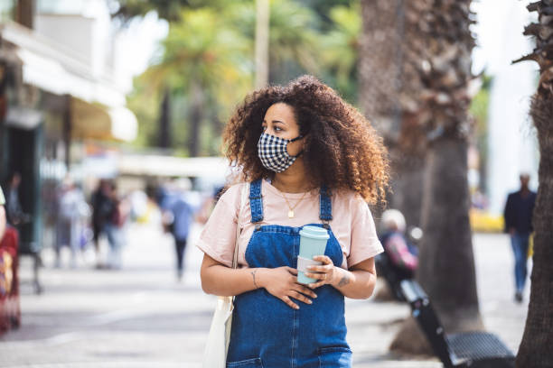 беременная женщина гуляет по городу в солнечный день, защищаясь с помощью ткани маски для лица - curley cup стоковые фото и изображения