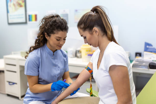 Preparation for blood test with pretty young woman by female doctor medical uniform on the table in white bright room. Nurse pierces the patient's arm vein with needle blank tube. stock photo