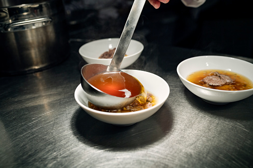 Chef carefully pouring a ladle full of wild consommé into a bowl that contains a delicate mix of carrots, leeks and celery a smoked egg yolk and some tender strips venison. Colour, horizontal with some copy space. Shot on location in a restaurant on the island of Møn in Denmark.
