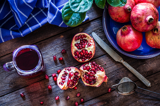 Top view of a rustic wooden table with a glass jar filled with pomegranate juice. Some pomegranates are in a blue plate and one is cut in half. A blue napkin is at the top-left corner. Some seeds are spilled on the table. Predominant colors are red and blue. Low key DSRL studio photo taken with Canon EOS 5D Mk II and Canon EF 100mm f/2.8L Macro IS USM