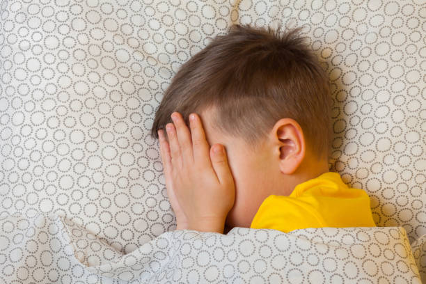 A preschool boy of 5 years lies in bed and covers his eyes with fear stock photo