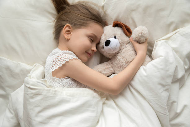 Preschooler girl hug teddy sleeping peacefully in cozy bed Smiling cute little girl hug teddy bear sleep peacefully in white bed relaxing on soft pillow, calm preschooler child enjoy daydream with plush toy taking nap under warm fluffy duvet at home fluffy stock pictures, royalty-free photos & images