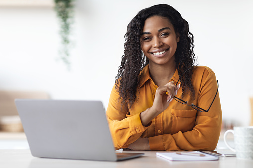 Pretty long-haired millennial african american woman freelancer using laptop at home, black lady sitting at table at kitchen, holding eyeglasses and smiling at camera, copy space. Remote job concept