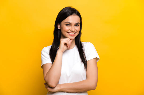 A pretty caucasian brunette girl dressed in a white basic t-shirt  stands on isolated yellow background, looks directly into the camera with a friendly smile stock photo